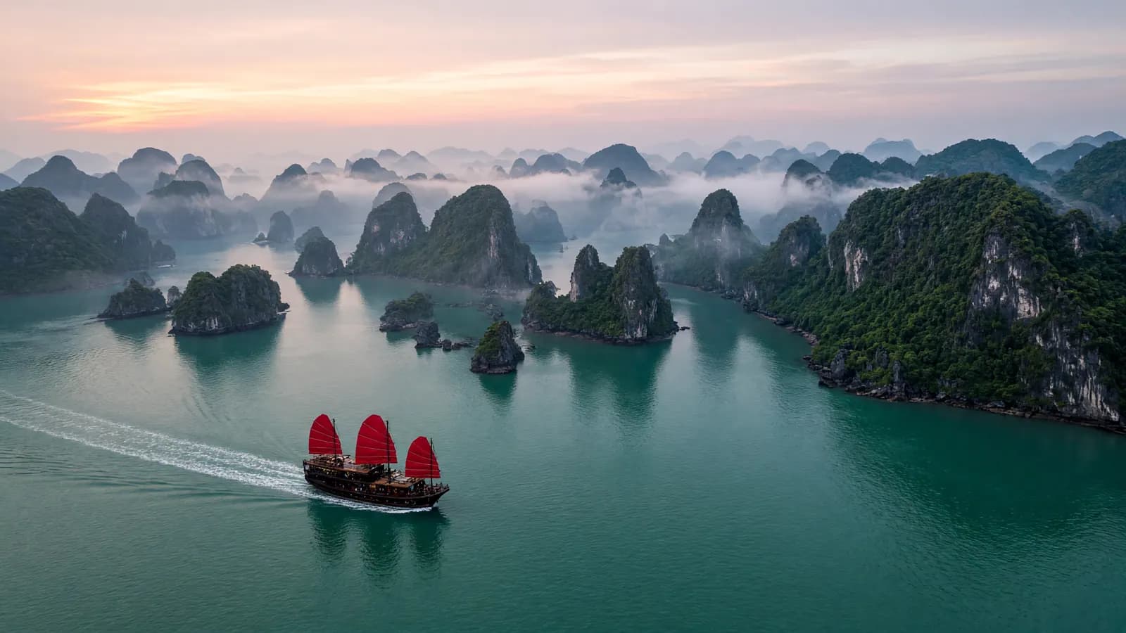 Vịnh Hạ Long lúc bình minh nhìn từ trên cao, thuyền buồm đỏ băng qua mặt nước.